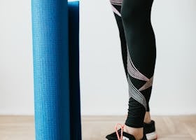 A woman in leggings and sneakers standing with a yoga mat indoors.