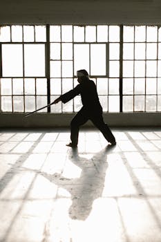 Silhouette of martial artist practicing with sword in a sunlit room, demonstrating skill and focus.
